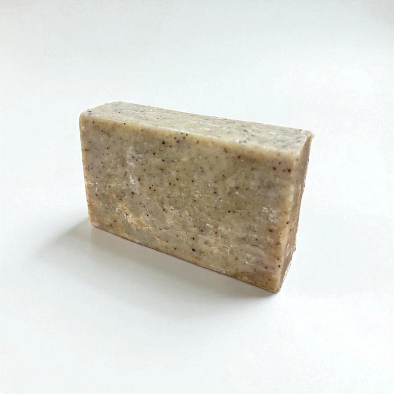 Pain rectangulaire gris et grainé de savon exfoliant à la nigelle sur fond blanc